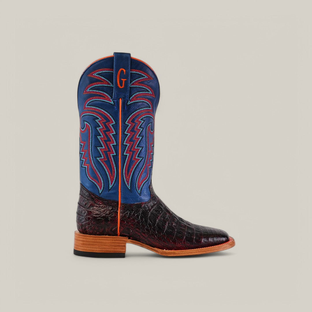 A handcrafted cowboy boot, similar to the Exotic Caiman Belly - Black Cherry - Square Toe, with a textured dark brown exotic leather lower and vibrant blue upper. It features red and blue stitching, an orange sole with low heel, and an embroidered G at the top.