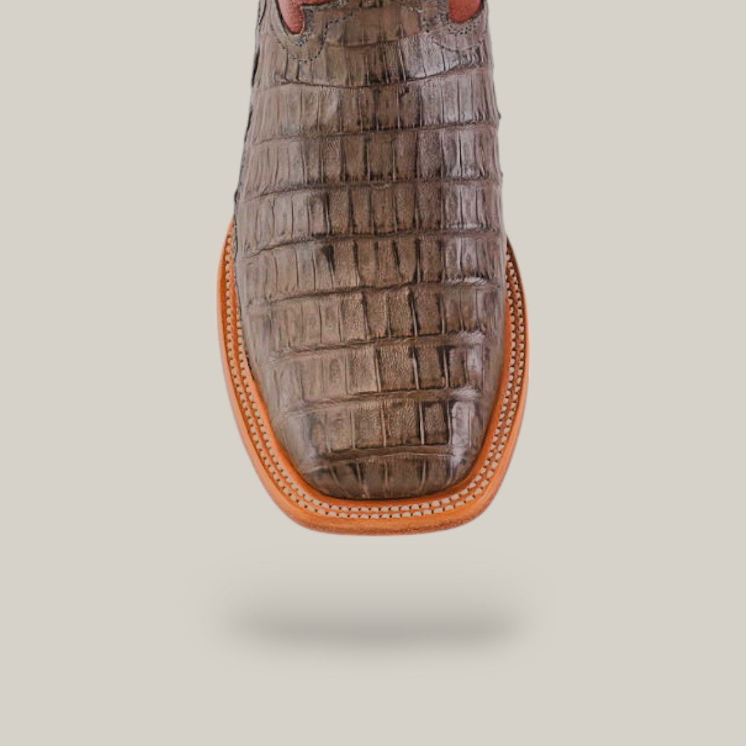 A close-up of the Exotic Caiman Belly Tail - Gray - Square Toe boot showcases its crocodile-patterned upper, reminiscent of caiman tail boots. It features a square toe and a contrasting tan leather sole with detailed stitching, all set against a plain, neutral background to highlight the Western style.