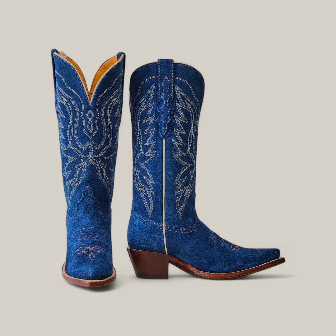 The Miranda Suede Blue Jeans boots showcase pointed toes and a sturdy leather sole, accented by intricate white and light blue stitching on the shaft and toe, with a plain beige backdrop.