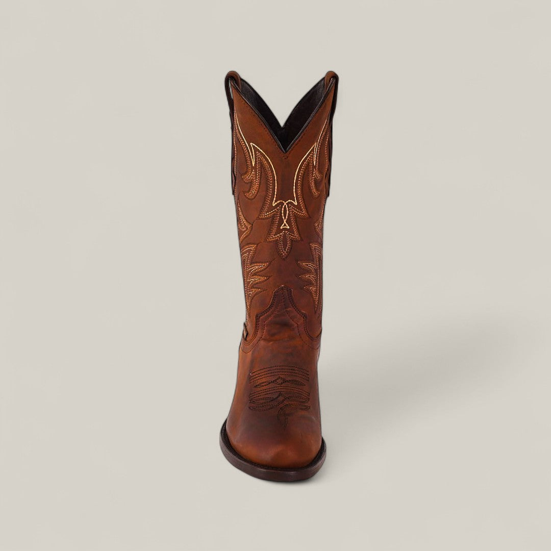 The Dutton - Brick - Roper Toe brown leather cowboy boot from the Last Call collection stands upright on a light beige background, with its cowhide toe pointed toward the viewer to highlight the decorative stitching.