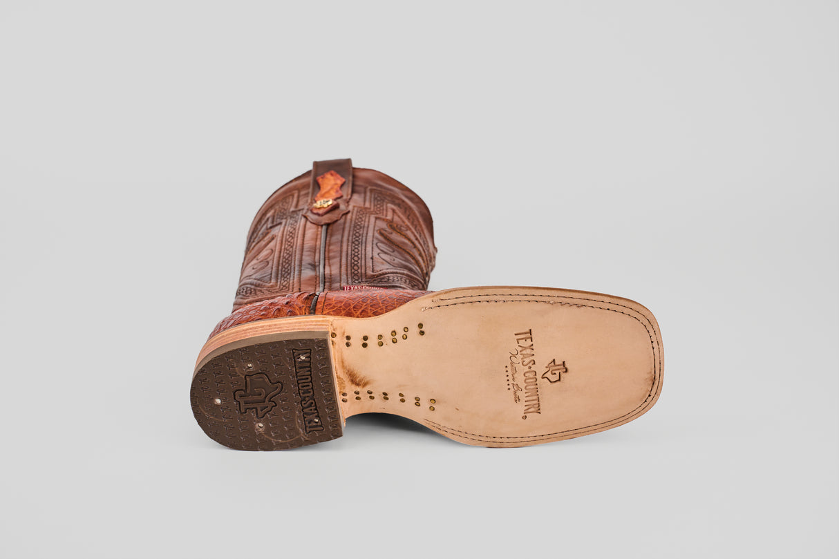 A single Exotic Caiman Belly - Brandy - Square Toe boot rests on its side, showing textured brown caiman leather and the TEXAS COUNTRY logo stamped on the light-colored sole against a plain light gray background.