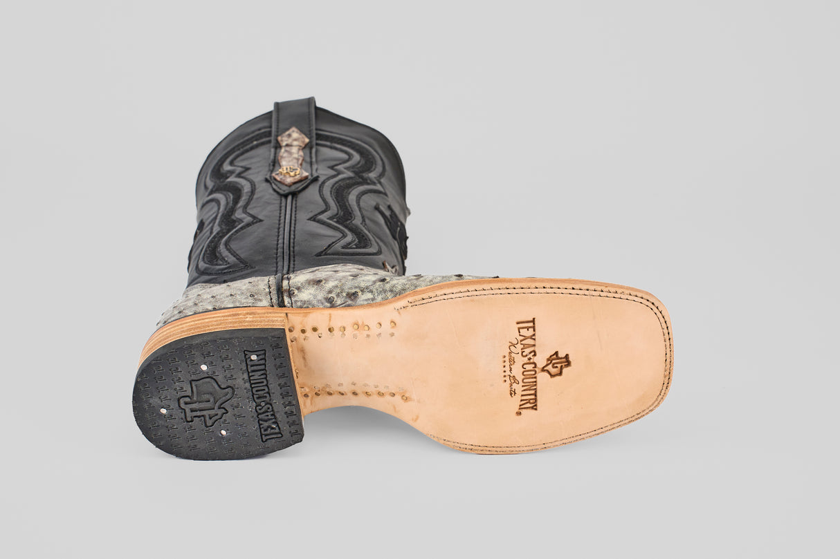 A cowboy boot from Exotic Full Quilt Ostrich - Nicotine - Square Toe lies on its side, showcasing a tan sole with the Texanosmith logo, black leather upper with decorative stitching, and an exotic quilted ostrich pattern near the ankle.