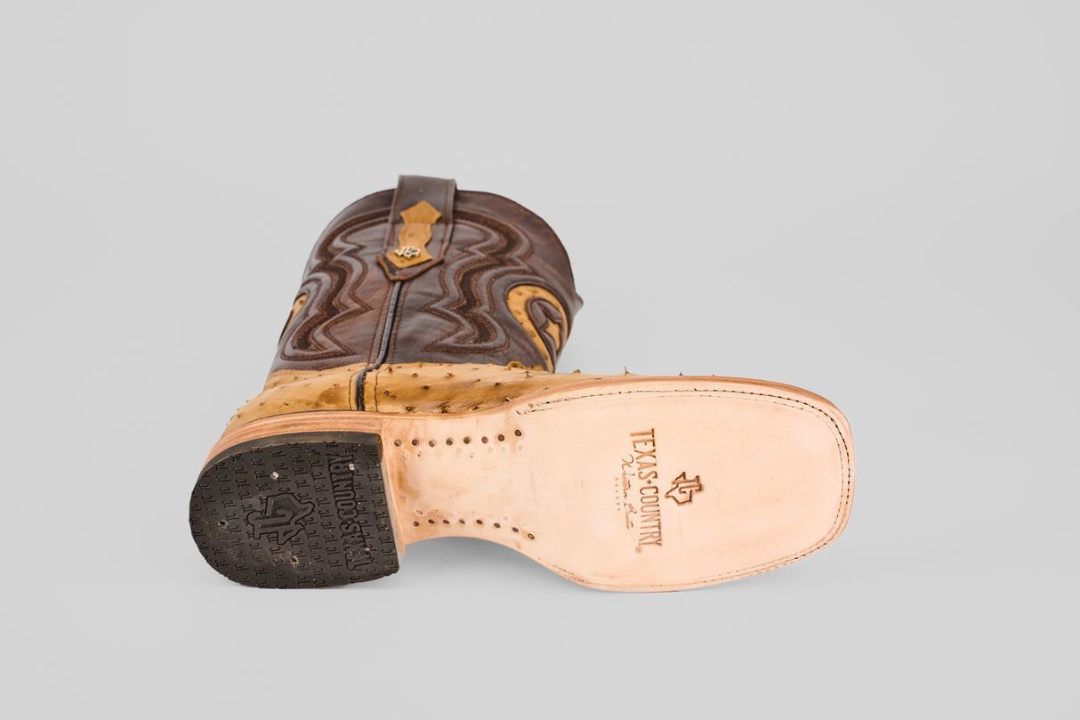 A single Exotic Full Quilt Ostrich - Fawn - Square Toe boot lies on its side, showing a light leather sole with the Texas Country logo, dark textured heel, and brown and tan decorative stitched leather.
