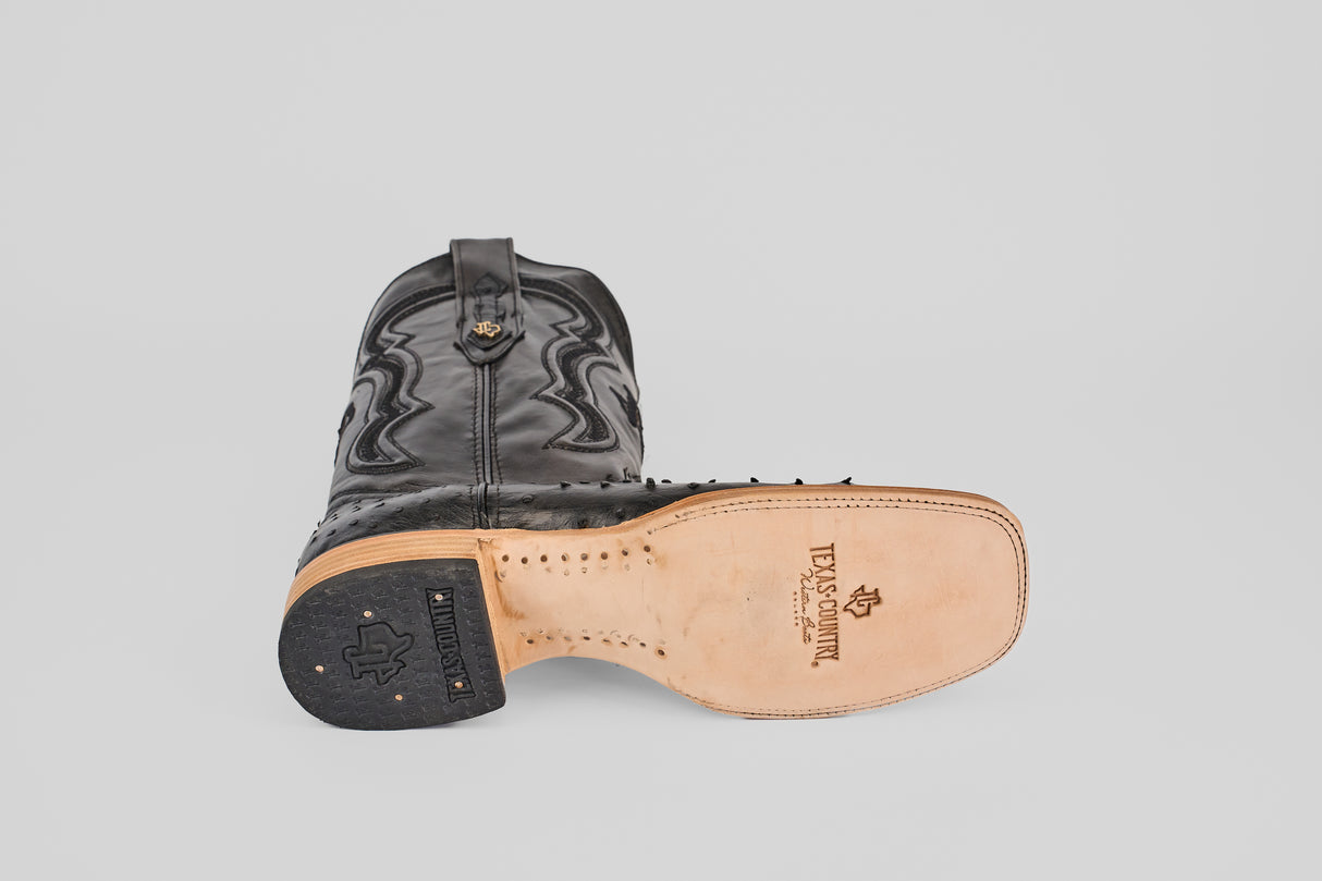 The Austin - Black - Square Toe boot lies on its side, highlighting detailed stitching and a tan leather sole with the Texas Country logo. Made from suave negro avestruz, it features a black heel adorned with small brass nails.