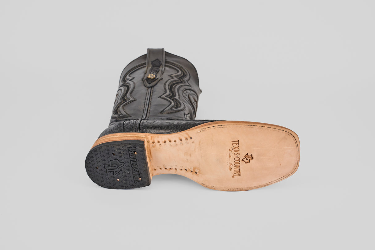 The Austin Leg - Black - Square Toe boot lies on its side, displaying its tan leather sole with the Texas Country logo. It features a square toe, decorative stitching, and a black rubber heel.