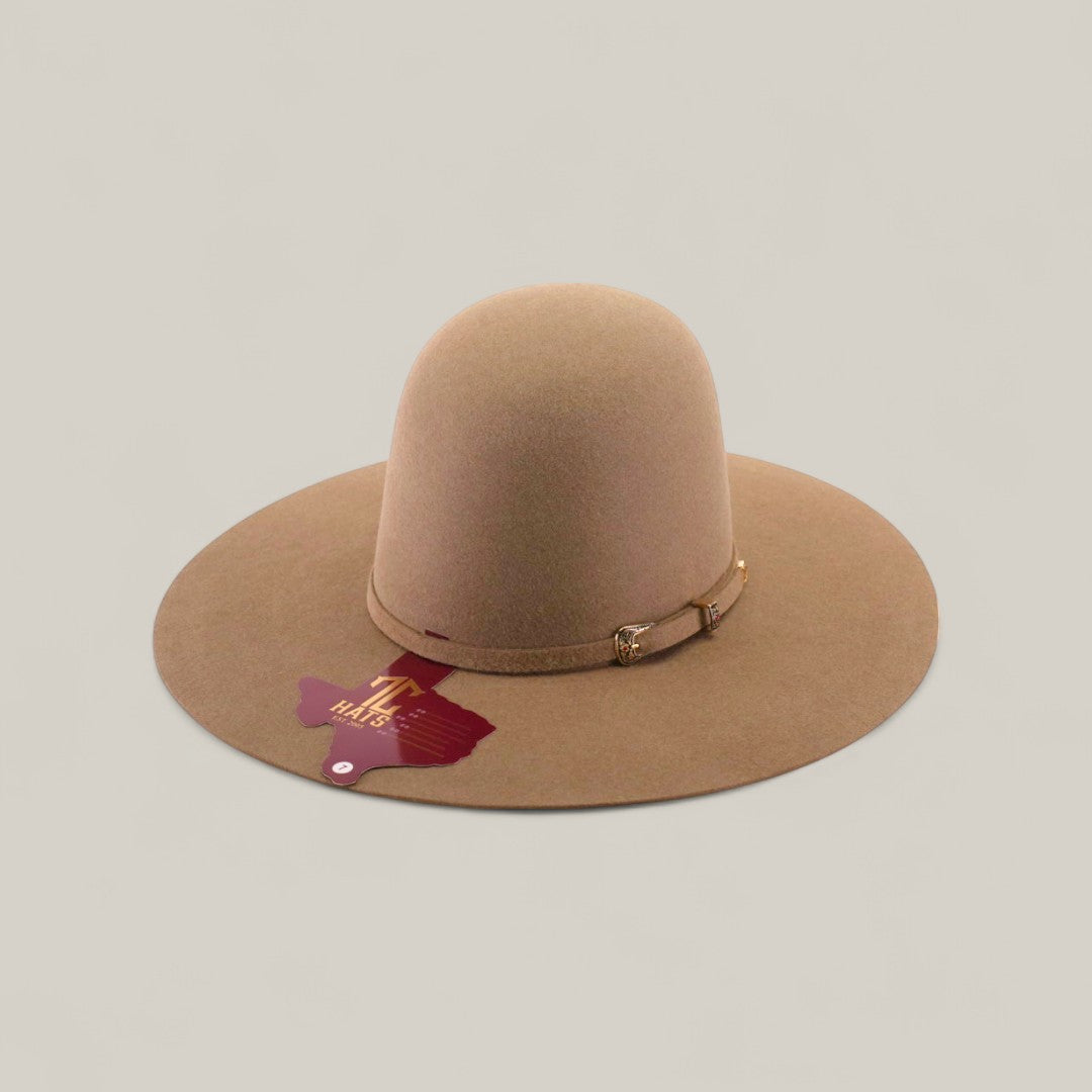The 10x - Natural - TC Hats is a wide-brimmed tan felt cowboy hat with a decorative band and buckle, shown on a light background. A maroon Texas-shaped tag hangs from the brim.