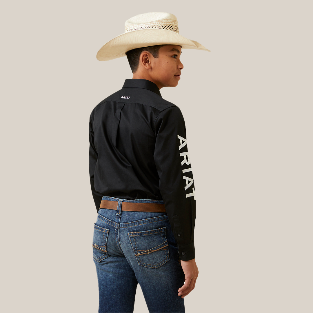 A young boy stands sideways in an Ariat Boys Black & White Team Logo Embroidered Long Sleeve Western Shirt (10045426), featuring a logo and white lettering on the sleeve and back. Hes paired it with blue jeans, a brown belt, and a wide-brimmed white cowboy hat, looking left against a plain white background.