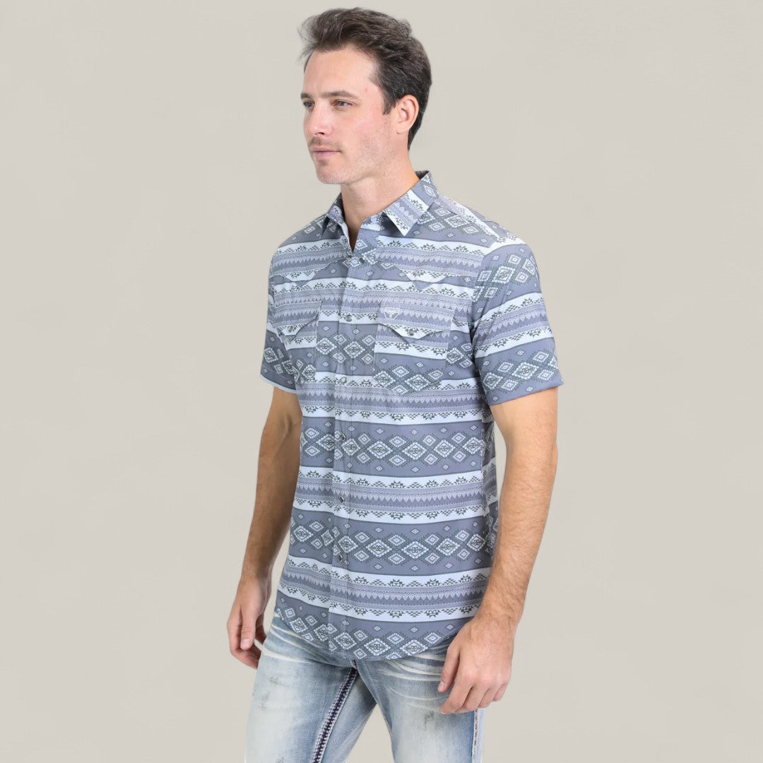A man with light skin and dark hair models the Mens Performance Western Short Sleeve Aztec Print Grey Shirt (PGW10649) featuring a blue and white geometric pattern. He stands against a plain beige background wearing light-wash jeans.