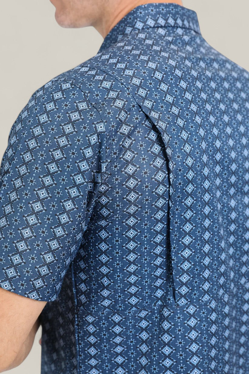 A close-up shows a person wearing the Mens Performance Western Short Sleeve Aztec Print Navy Shirt (PGS10659), featuring a bold geometric diamond and Aztec pattern. Only the upper body and part of the face are visible.