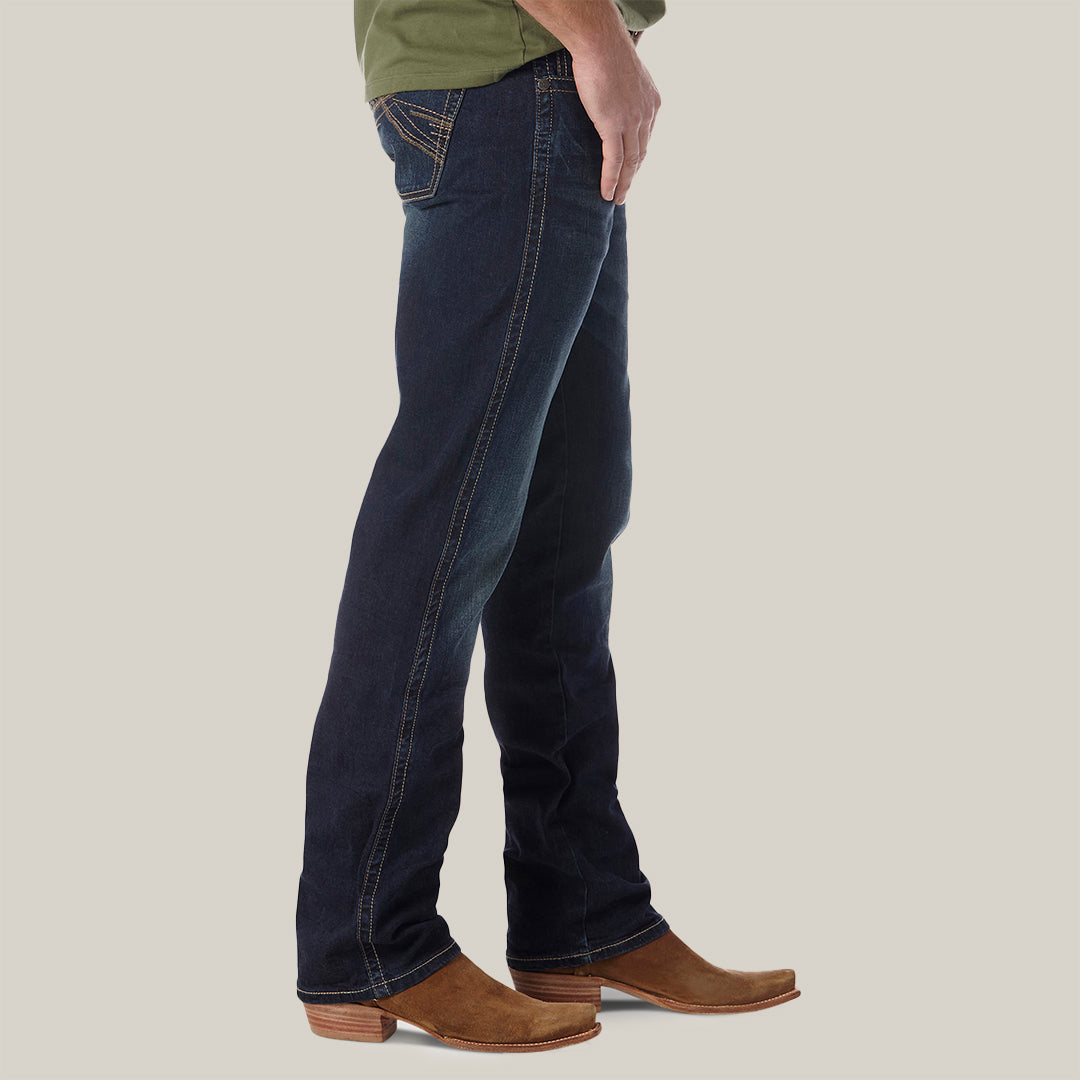 Side view of a person wearing Mens Wrangler 20X No. 42 Vintage Bootcut Jeans (42MWXRD) in dark blue with light brown cowboy boots, against a white background. The jeans, featuring premium contrast stitching on the back pocket, are visible from the chest down.