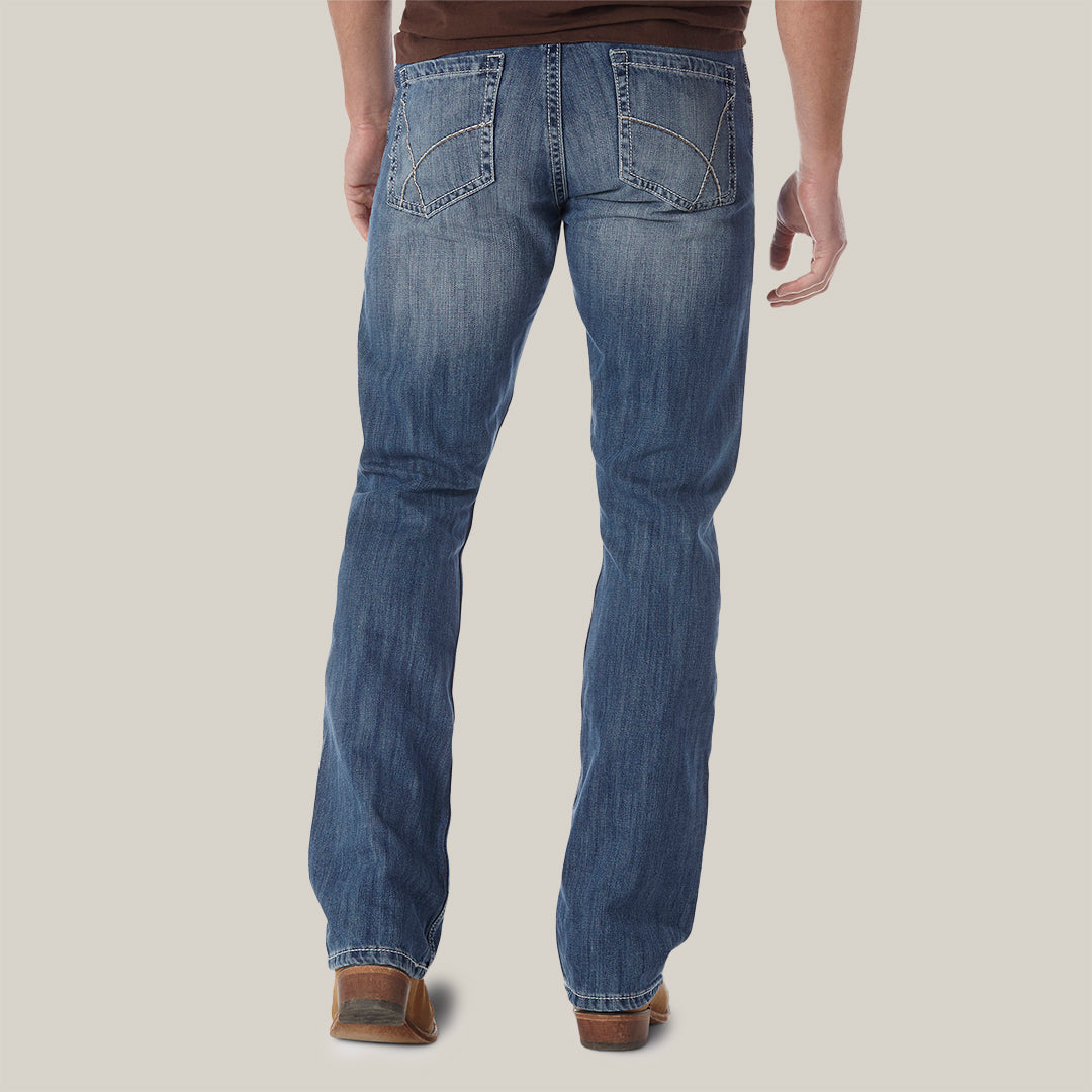 Back view of a person wearing Wrangler 20X No. 42 Vintage Bootcut Jeans (1042MWXLB) and a brown shirt, standing against a plain backdrop. The jeans have decorative stitching on the back pockets and are paired well with brown shoes.