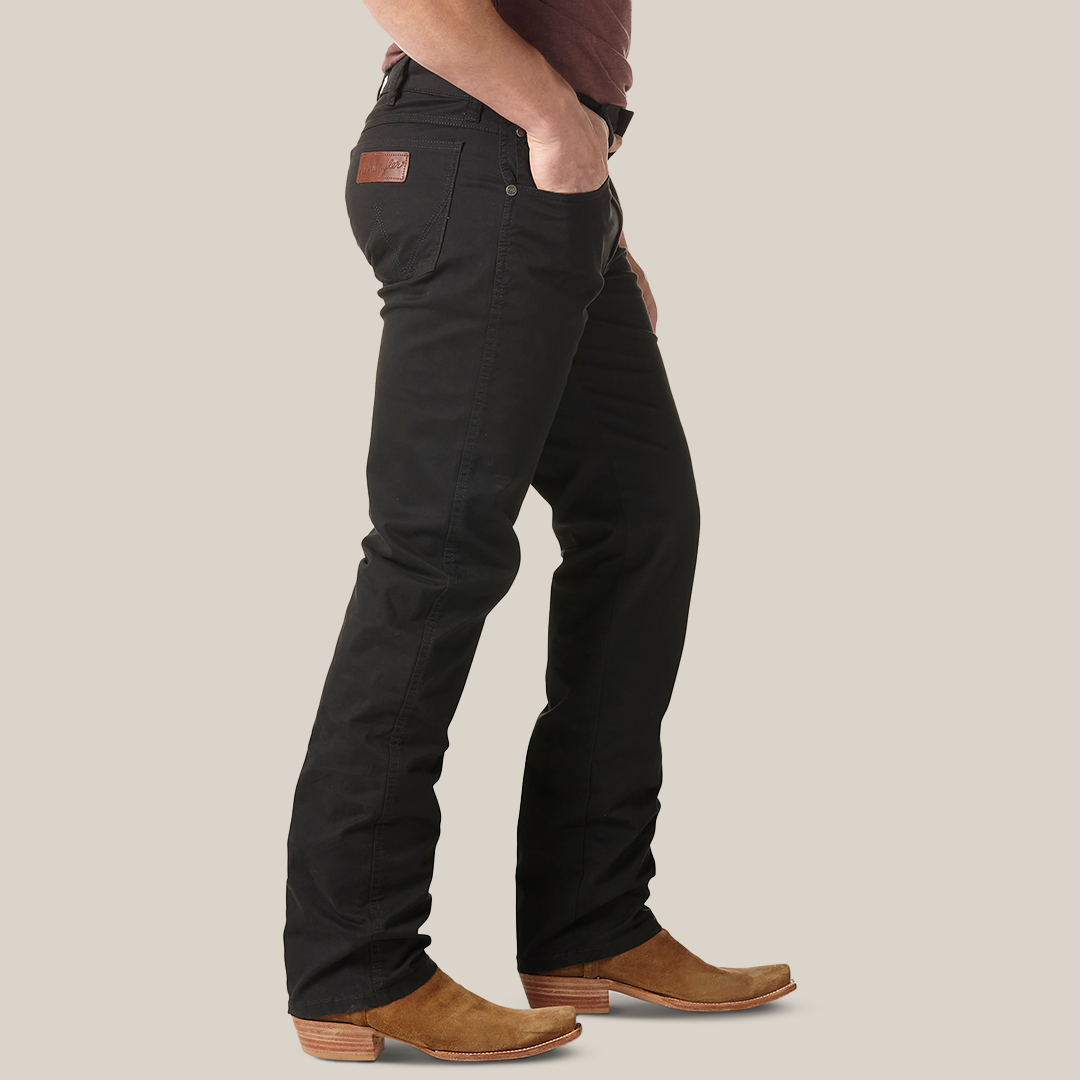 A person in a maroon shirt and Mens Wrangler Retro Slim Fit Straight Leg Pant - 88MWZBK with tan cowboy boots stands sideways, hand in pocket, against a light gray backdrop, highlighting the premium denims stylish appeal.