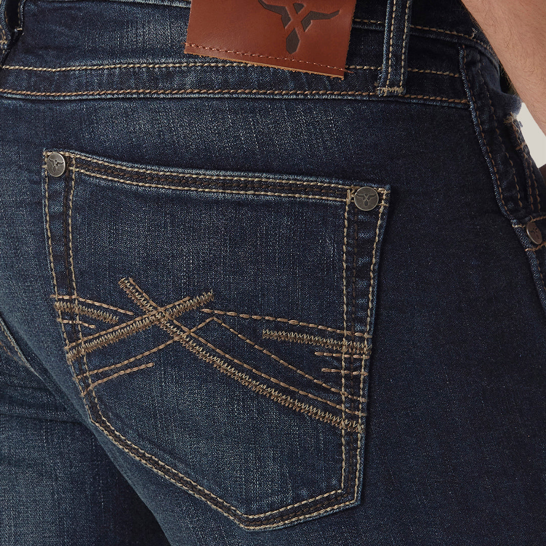 A close-up shows a person in Mens Wrangler 20X No. 42 Vintage Bootcut Jeans. These dark blue slim fit jeans boast tan stitching with intricate patterns on the right back pocket and a brown leather logo patch on the waistband, perfectly complementing a green shirt.