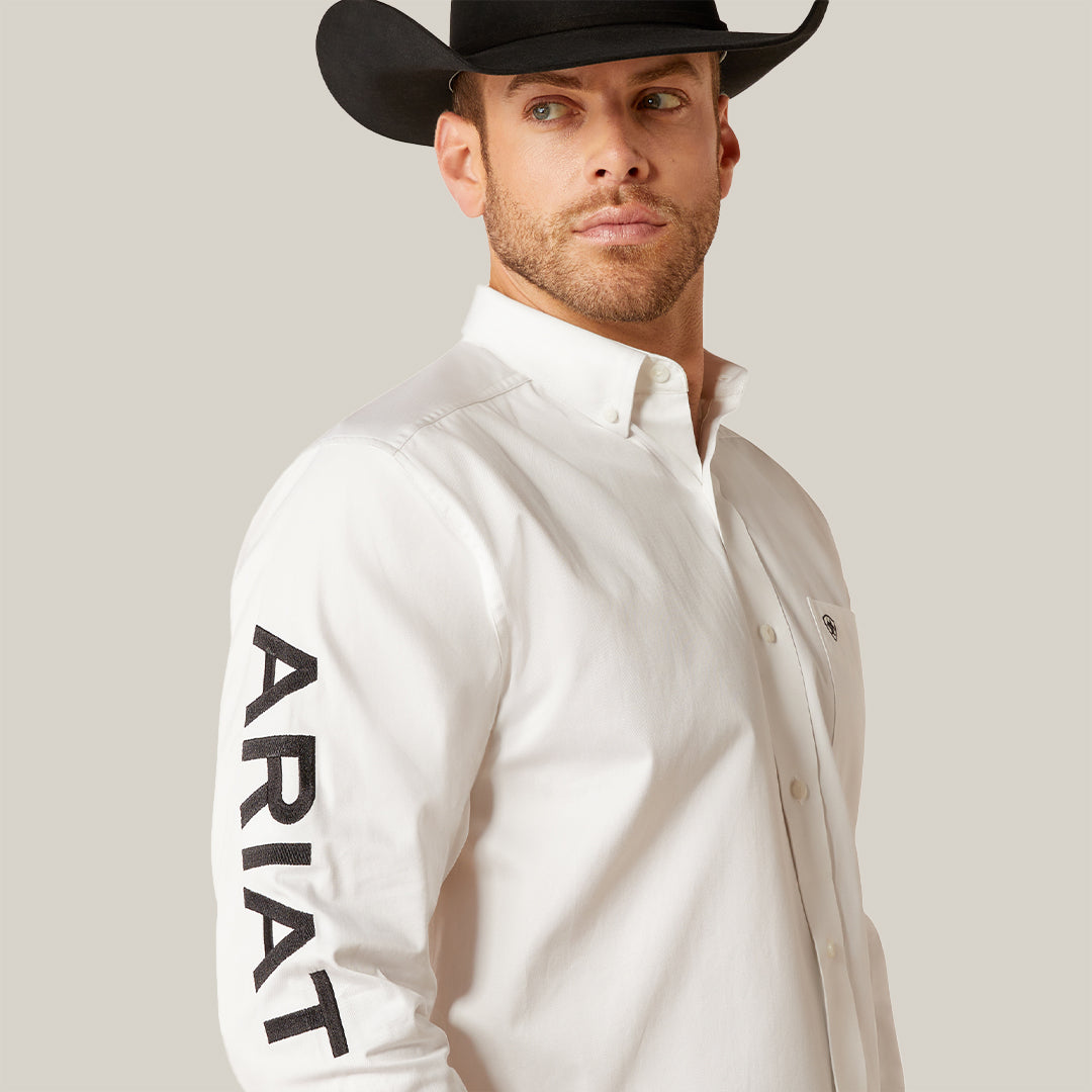 A man in a white ARIAT Team Logo Twill Classic Fit Shirt (10046825) glances left. His black cowboy hat pairs well with the subtle chest pocket logo and embroidered details, enhancing the timeless appeal of the shirts classic fit and plain white background.