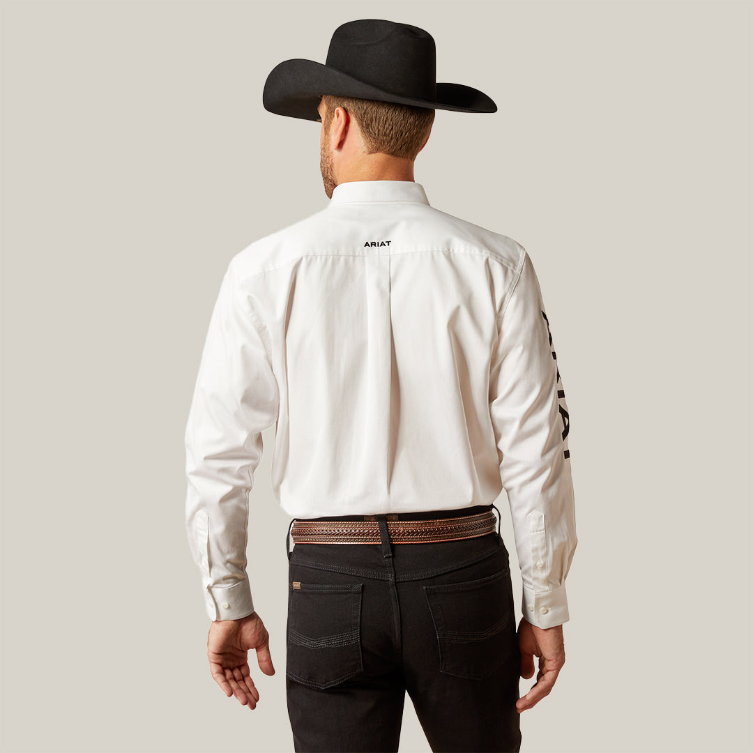 A man in a Team Logo Twill Classic Fit Shirt White (10046825) with embroidered details pairs it with a black cowboy hat and jeans, fastened by a brown leather belt. The back view reveals a logo on the shirts upper back and text on the left sleeve.