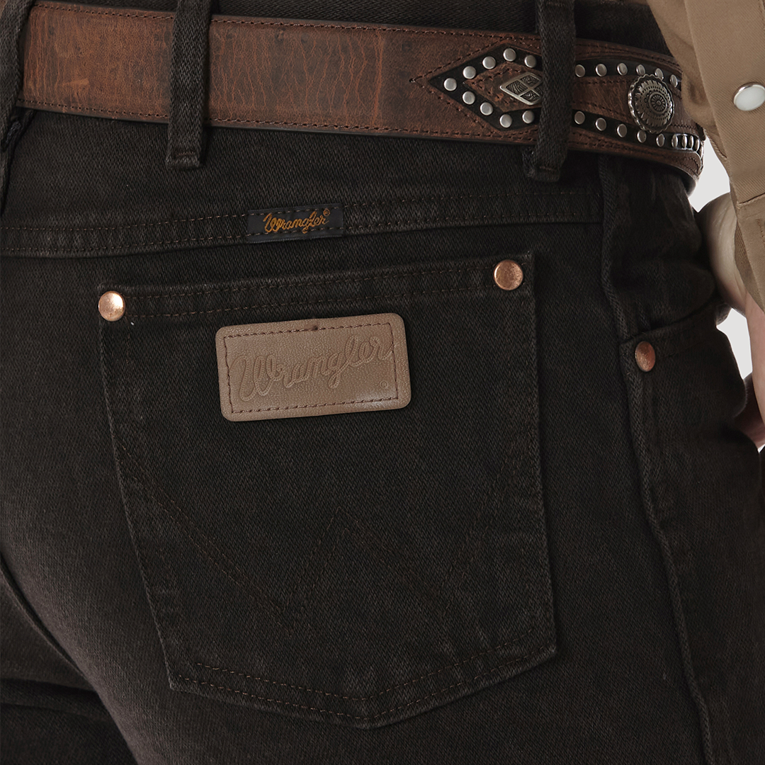 Close-up of a person in Wrangler Cowboy Cut Slim Fit Jean - 0936KCL, dark denim with a leather belt featuring silver details. The tan shirt is partially visible, but the iconic Wrangler label on the back pocket remains the focus.