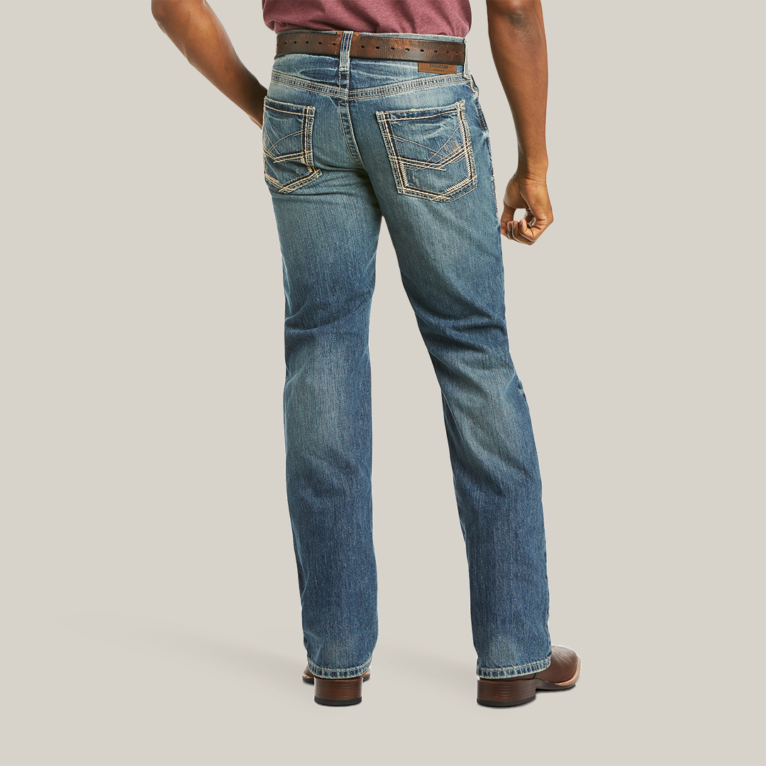 View of a person wearing the M5 Slim Gambler Stackable Straight Leg Jeans (10012703) with stitched back pockets, a brown belt, and brown shoes. The durable denim enhances their silhouette as they stand confidently with their left hand on their hip against a white background.