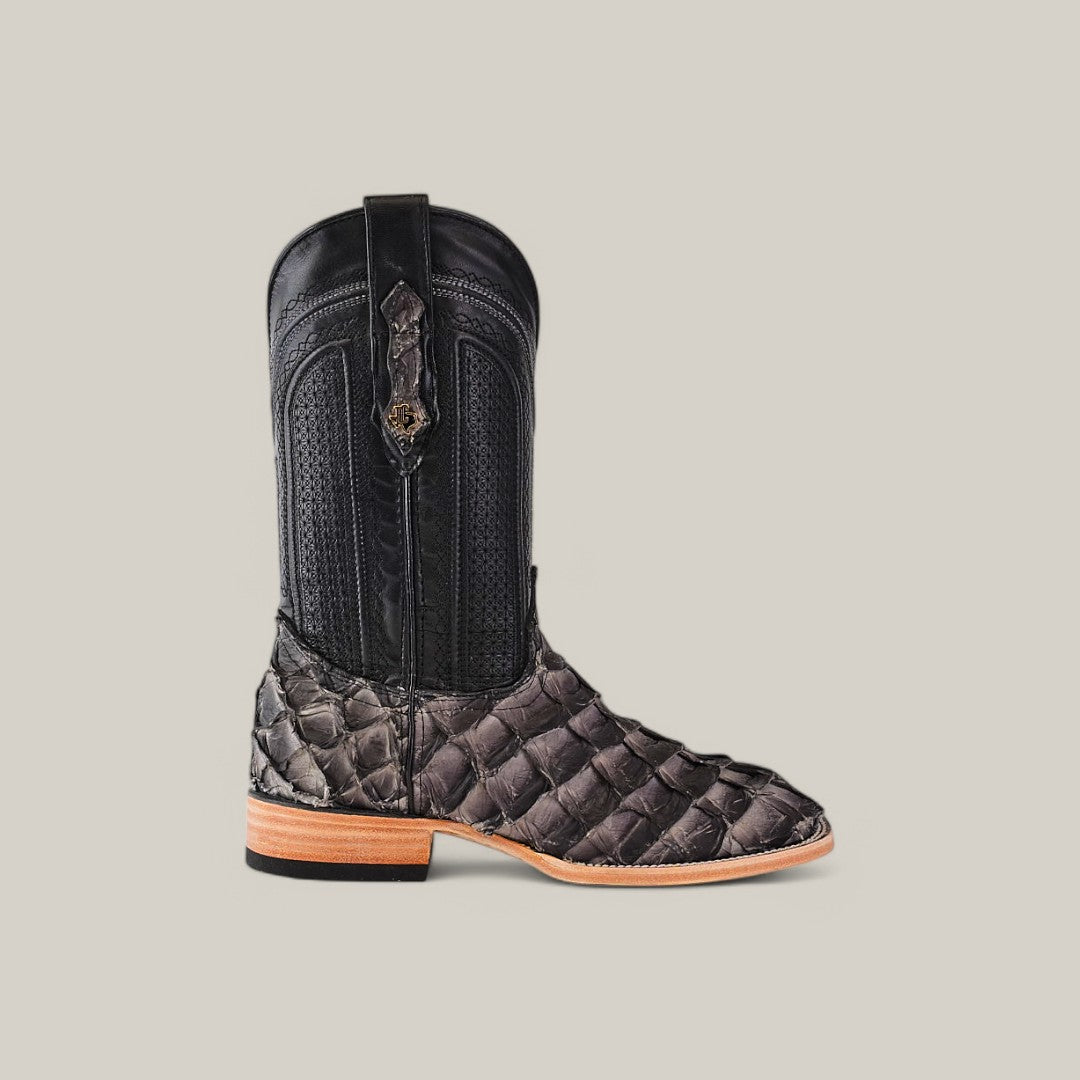 The Exotic Pirarucu Fish Gray boot features a single cowboy boot in dark textured leather with intricate stitching, a square toe, and wooden heel. The detailed upper section includes a strap and loop for easy wear. Crafted from sustainable materials, its set against a solid light gray background.