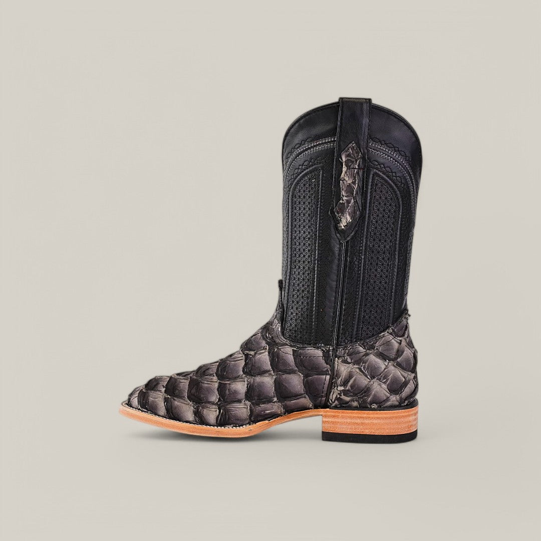 The Exotic Pirarucu Fish cowboy boot features a square toe, a black textured upper, and a brown scaly lower part with a smooth wooden heel on a plain light background.