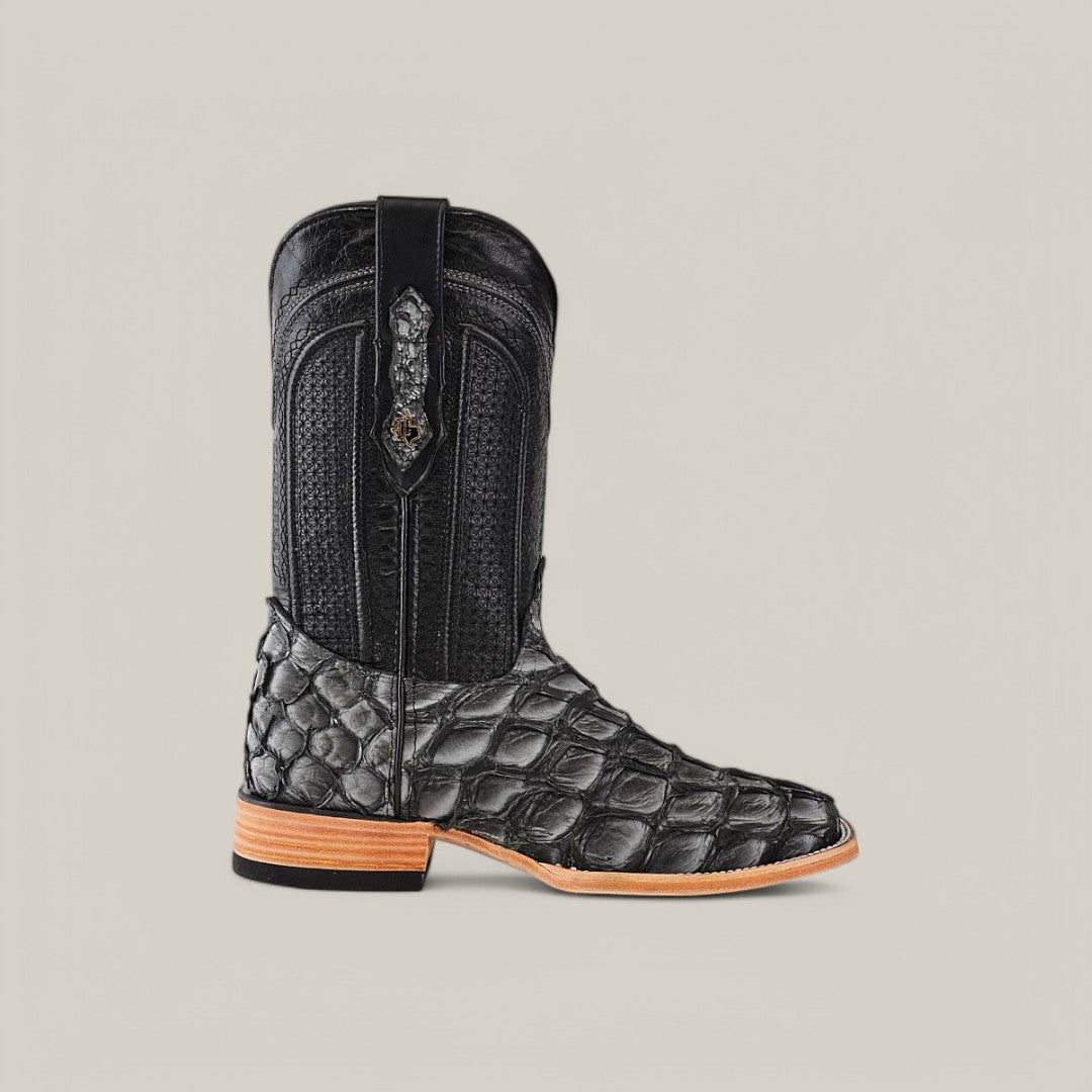Set against a neutral backdrop, the Exotic Pirarucu Fish - Newspaper - Square Toe boot flaunts black textured exotic leather with intricate stitching, a decorative pull strap, and a modern square toe, complemented by a natural-colored sole.