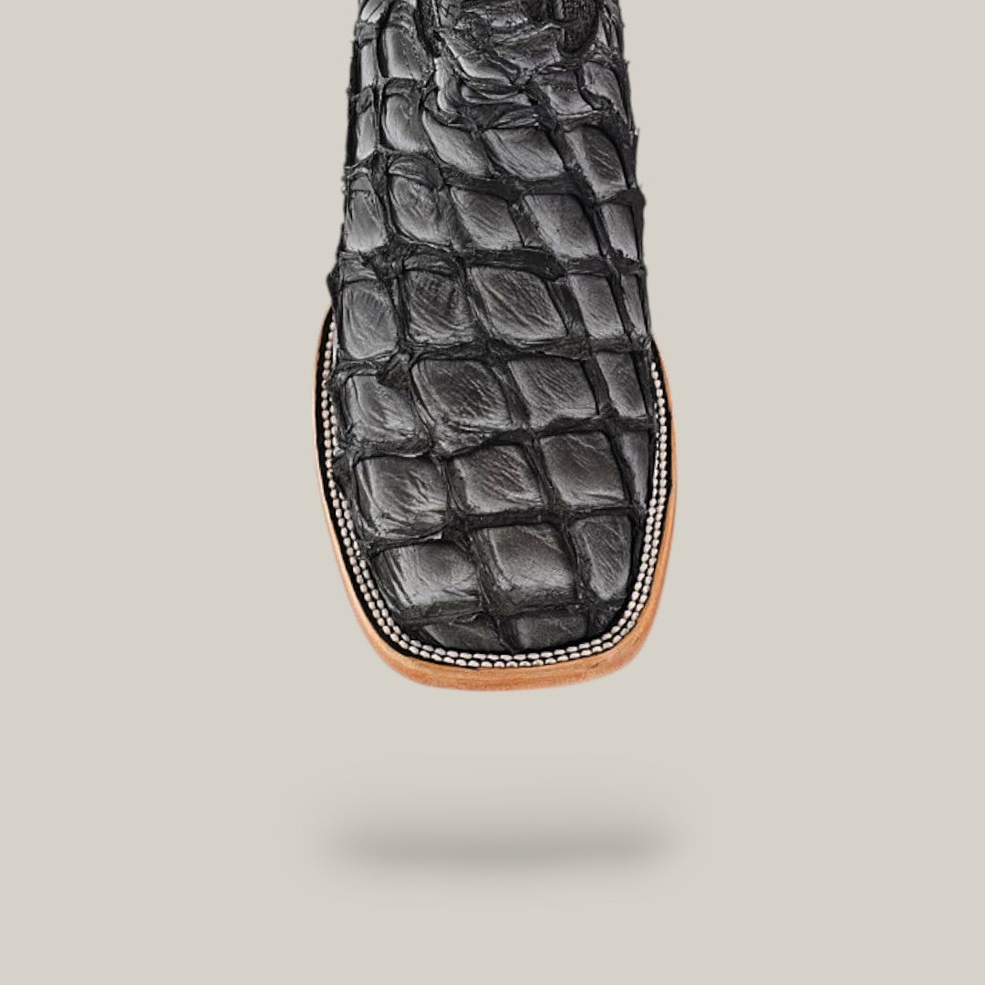 Close-up of a black, square-toed boot from the Exotic Pirarucu Fish - Newspaper collection, featuring a textured scaly look and contrasting brown sole against a light background.