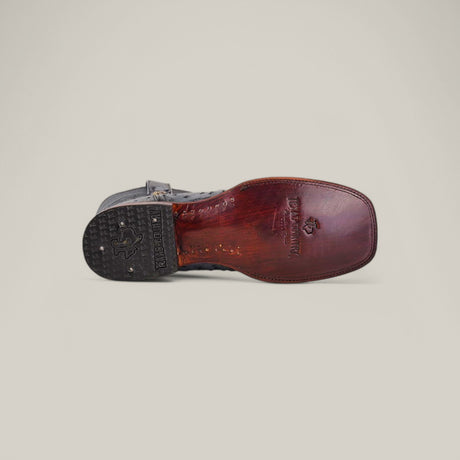 Bottom view of the Exotic Full Quilt Ostrich - Safari Gray - Square Toe boot with a red-brown leather sole featuring engraved text and logo. The black heel has a decorative rubber grip pad, offering sophistication to any ensemble against the plain light gray background.