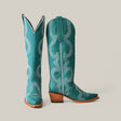 A pair of Olimpia Green Teel cowboy boots features intricate white stitching and patterns. One boot faces forward, while the angled snip-toe design showcases the premium materials and stacked wooden heel, all set against a plain neutral background.