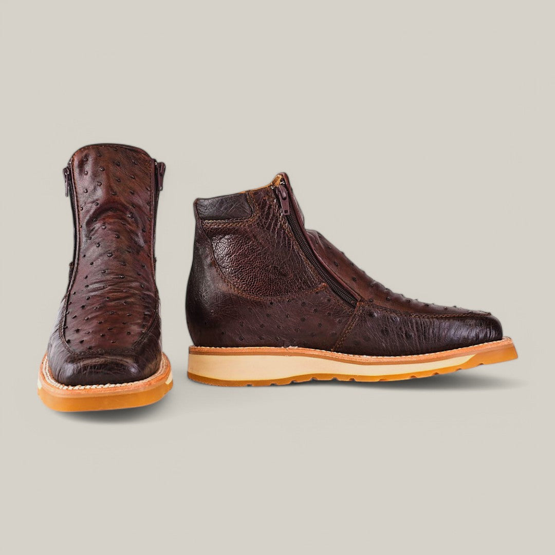 The Exotic Ostrich ankle boot in dark brown features textured ostrich leather with light tan soles. One boot is upright showing the side, while the other tilts forward displaying the front, both with visible side zippers against a plain, light gray background that highlights this luxury footwear.