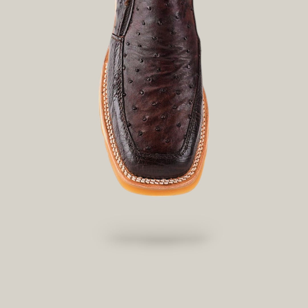 The Exotic Ostrich - Dark Brown Ankle Boot is showcased against a light gray background, featuring dark brown ostrich leather, a square toe, detailed stitching, and a tan sole. Subtle shadows suggest its slight elevation.
