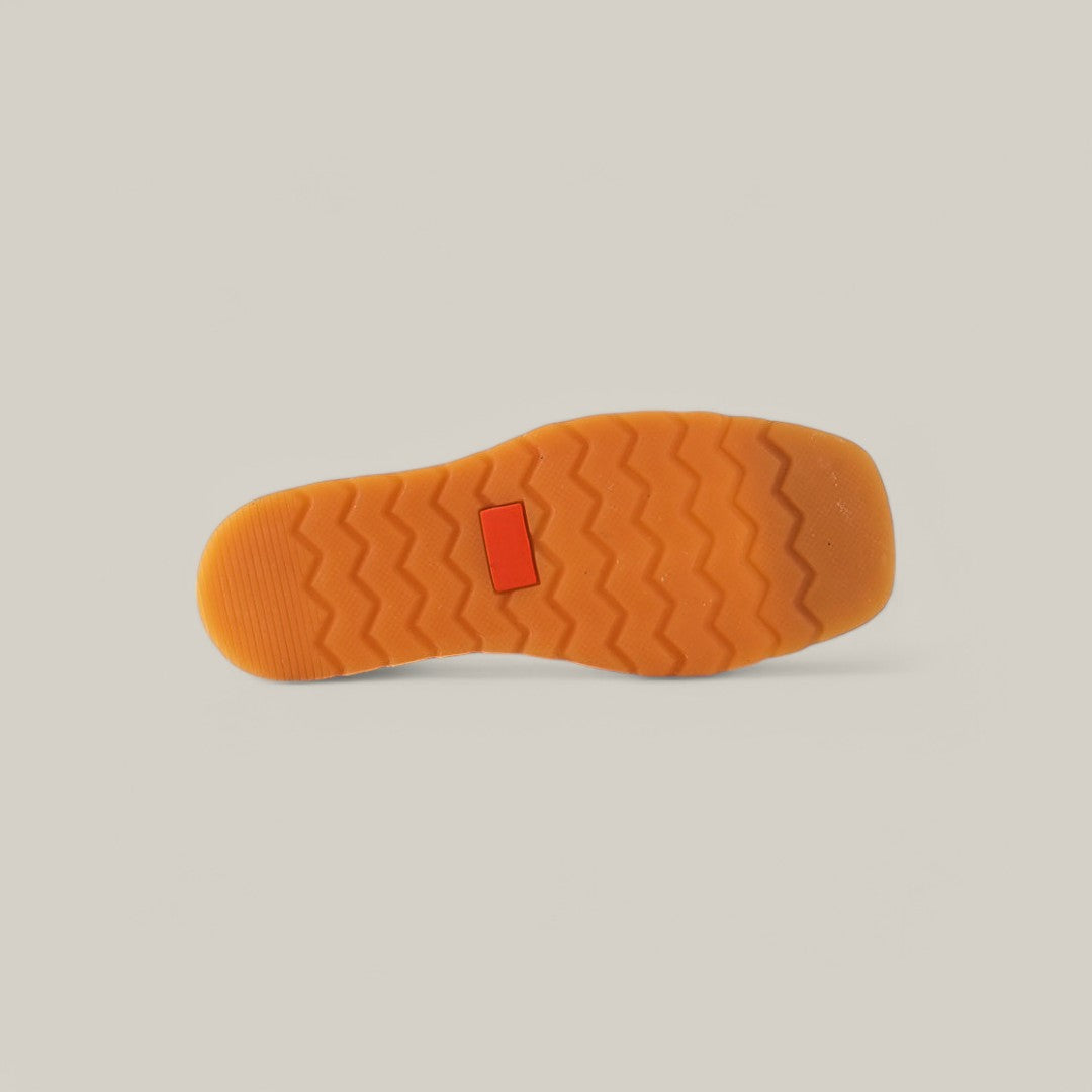 The image highlights the bottom of an orange sole with a zigzag pattern and a small red rectangle, epitomizing luxury. Set against a neutral beige background, it accentuates the unique design of this Exotic Ostrich Ankle Boot in Dark Brown.