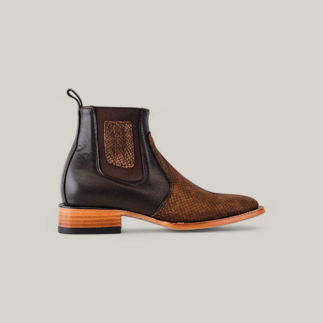 The Exotic Python - Tamarindo is a stylish two-toned ankle boot featuring a brown woven vamp with a dark Python-embossed leather upper, elastic side panels, and pull tab. Its set on a wooden sole with a low heel against a neutral backdrop.