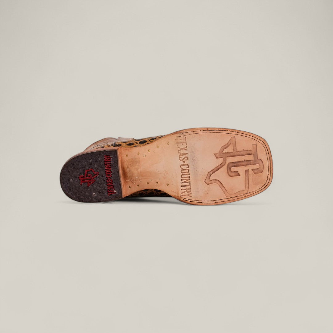 The Jonh - Orix - Square Toe boot features a flat tan leather sole stamped with “TEXAS COUNTRY” and a bold TC logo. Its dark heel displays a red Texas Country emblem set against a plain, light background.