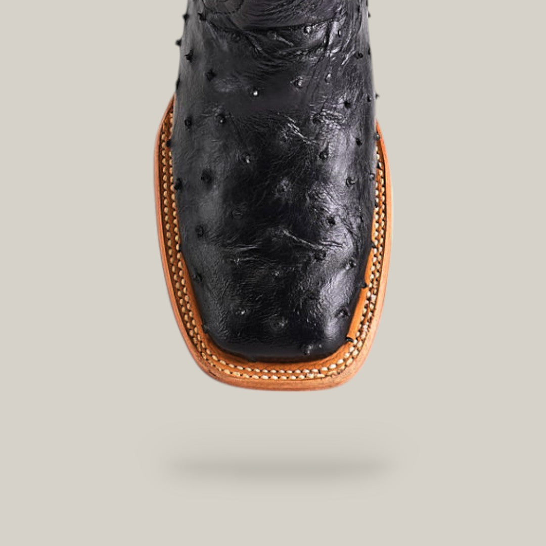 A close-up of the Exotic Full Quilt Ostrich - Black - Square Toe boot highlights its black ostrich leather, square toe, and tan stitched welt against a light gray background.