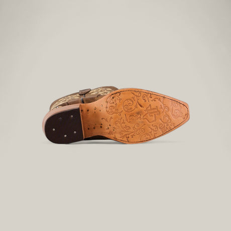 The Roxanne Crystals Tabaco boot lies on its side, displaying a sole with floral patterns and a logo. Its tall shaft and snip toe design stand out, while the dark brown heel with visible nails adds to its luxurious women’s style.