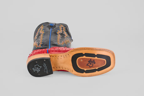 The Crowley - Red - Square Toe cowboy boot lies on its side, showing the leather and rubber sole. The upper features black and red leather with yellow decorative stitching and blue piping.