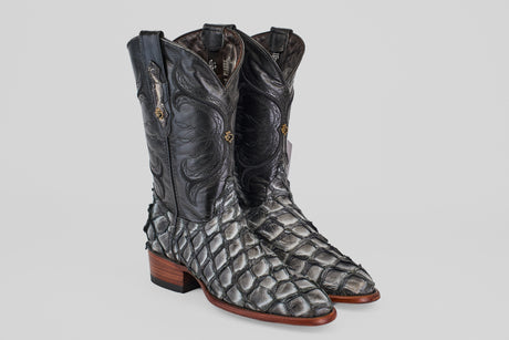The Conor - Newspaper - Roper Toe boots feature dark, intricately patterned uppers and silver-gray scaly Pirarucu fish leather feet, complemented by medium brown stacked heels and side stitching, set against a plain background.