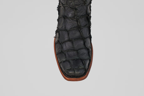 A close-up of the Conor - Matte Black - Narrow Square Toe boot shows its textured, fish leather-like pattern and brown sole, set against a plain gray background.