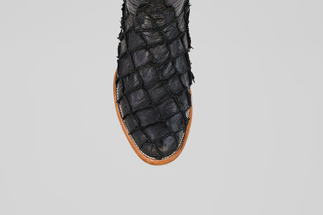 A close-up of the Conor - Matte Black - Roper Toe boot, featuring textured black pirarucu fish leather with a scale-like pattern and a brown sole, set against a plain light gray background.