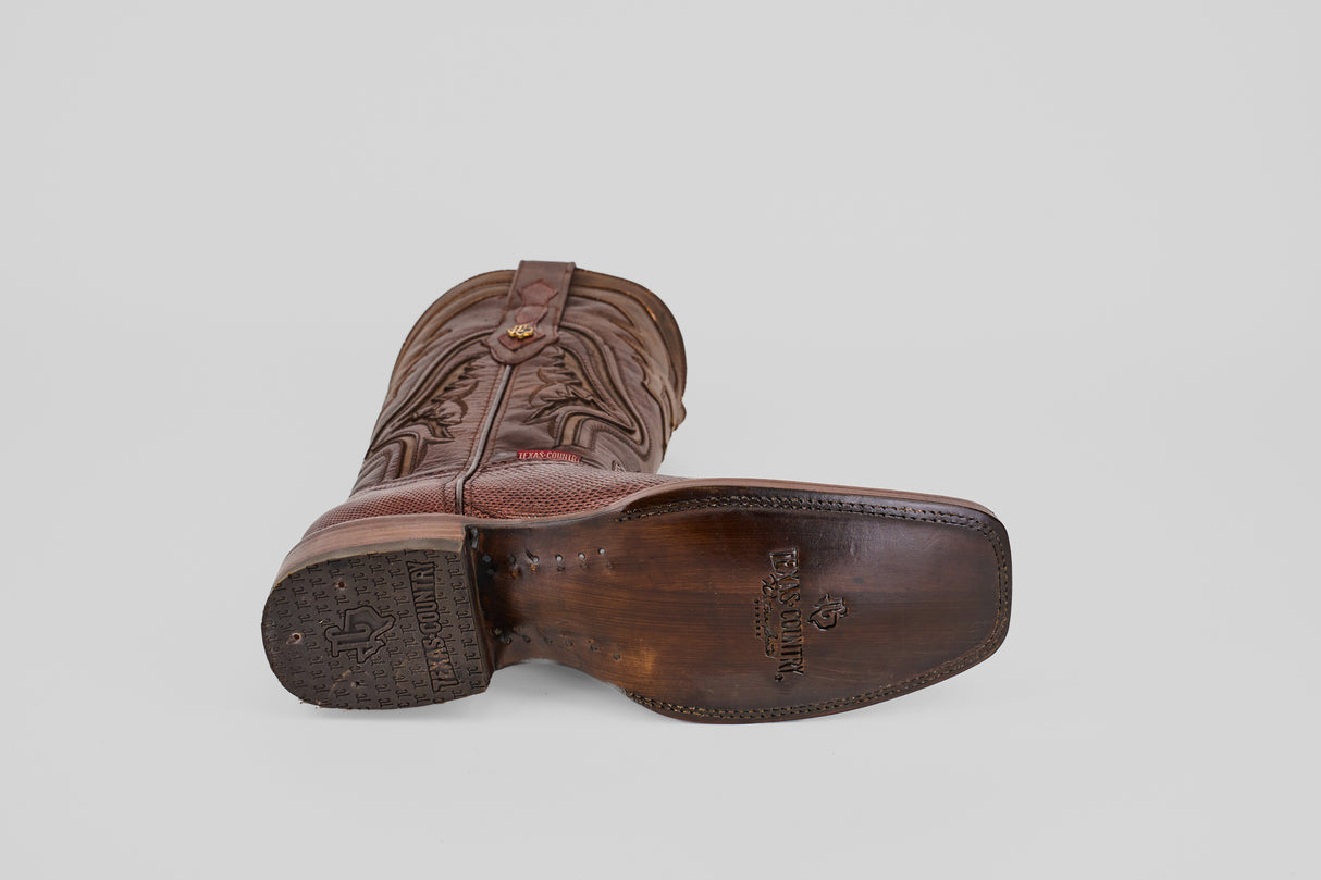 A single Blaze – Brown – Rodeo Toe cowboy boot lies on its side, showcasing the sole with Lucchese Bootmaker branding. Crafted from genuine lizard leather, it features decorative stitching, a pull strap, and a plain light gray background.