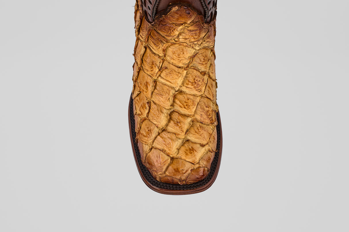 A close-up of the Conor Platinum - Buttercup - Square Toe (Herradura) cowboy boot highlights its unique pirarucu leather scale pattern and rich brown color, showcasing the premium exotic texture against a plain gray background.