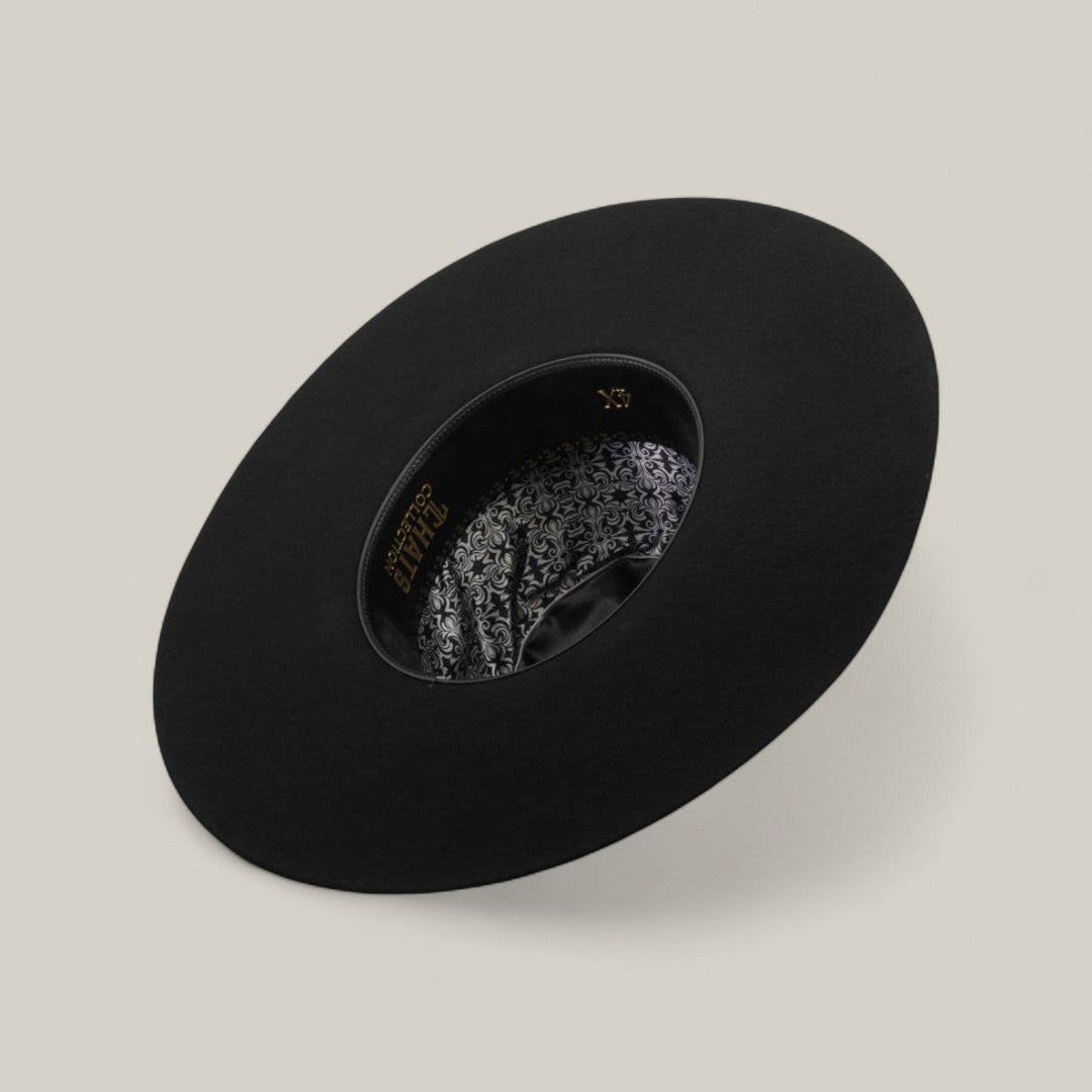The 4X - Black - TC Hat is displayed upside down, showing its ornate patterned lining with gold lettering. Its shapeable crown is set on a light beige surface.