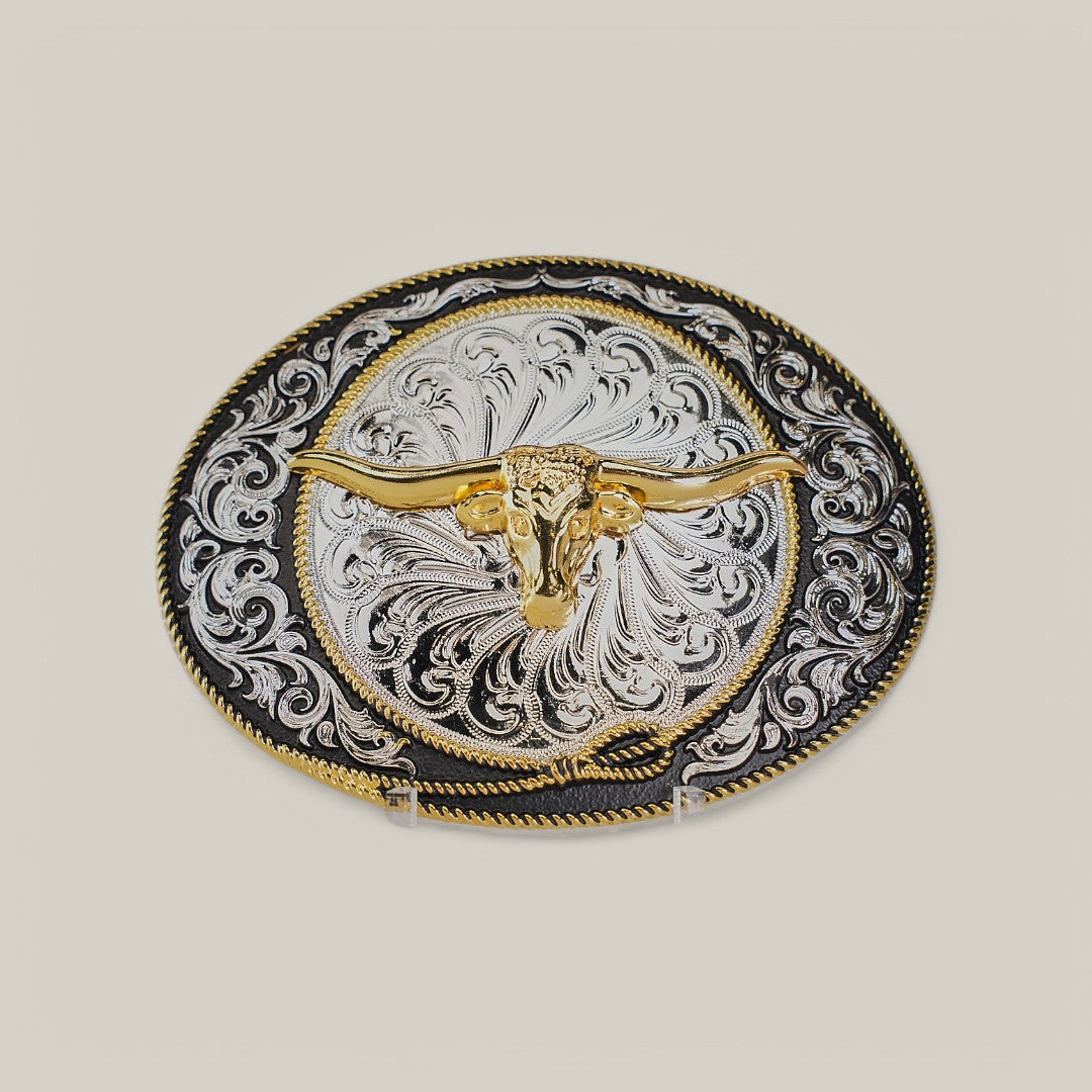 The Oval Gold Longhorn Buckle features an ornate design with a golden longhorn skull embossed at the center, surrounded by intricate alpaca silver filigree patterns on a contrasting dark background.