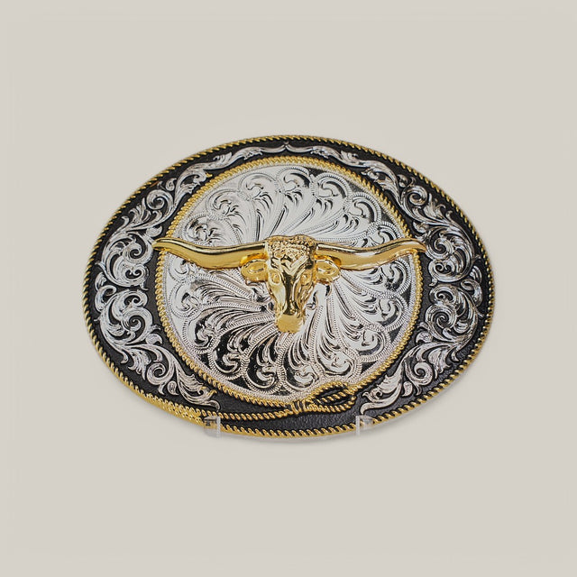 The Oval Gold Longhorn Buckle features an ornate design with a golden longhorn skull embossed at the center, surrounded by intricate alpaca silver filigree patterns on a contrasting dark background.