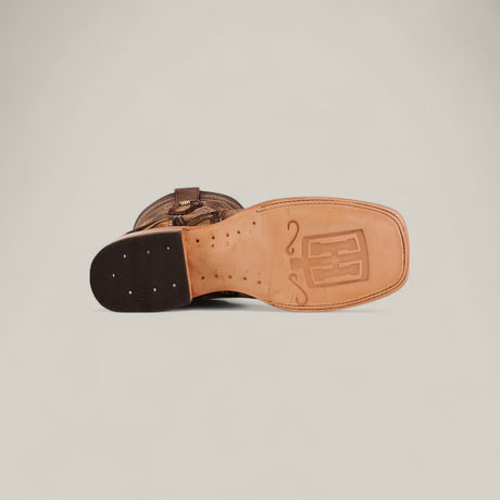 The Big Bass Pirarucu Print Rustic Camel - Square Toe cowboy boot is shown sole-up, revealing a light tan leather sole with a stitched heel, decorative logo and design etched on the bottom. The boots upper is partially visible.