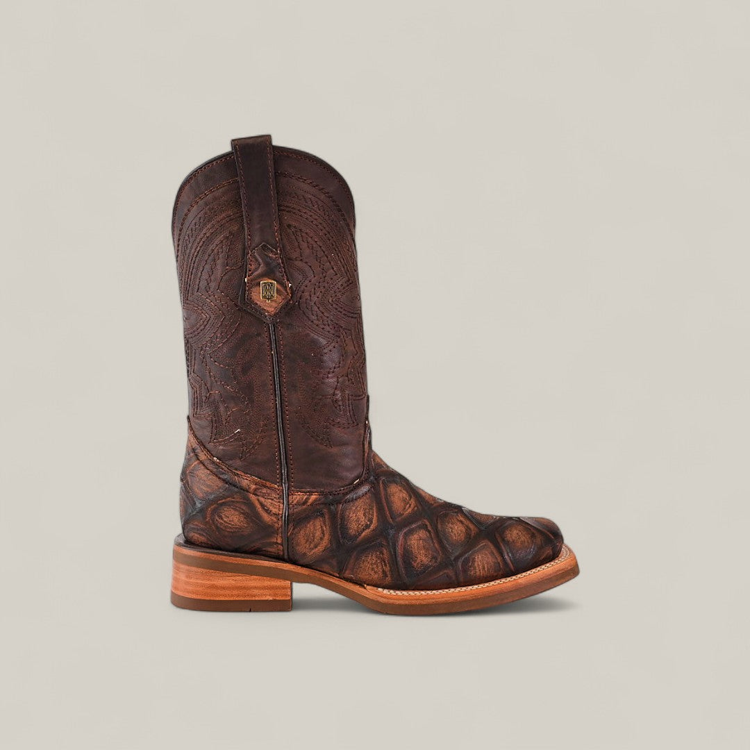 The Big Bass Pirarucu Print Rustic Brown - Rodeo Toe boot features a rustic brown finish, textured scale-like pattern on the lower part, detailed stitching on the shaft, and is displayed against a light beige background.