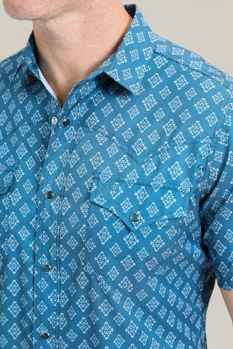 A man wears the Mens Performance Western Short Sleeve Aztec Print Blue Shirt (PGW10632) with a white geometric pattern, snap buttons, collar, and two chest pockets. Only his lower face and upper torso are visible.