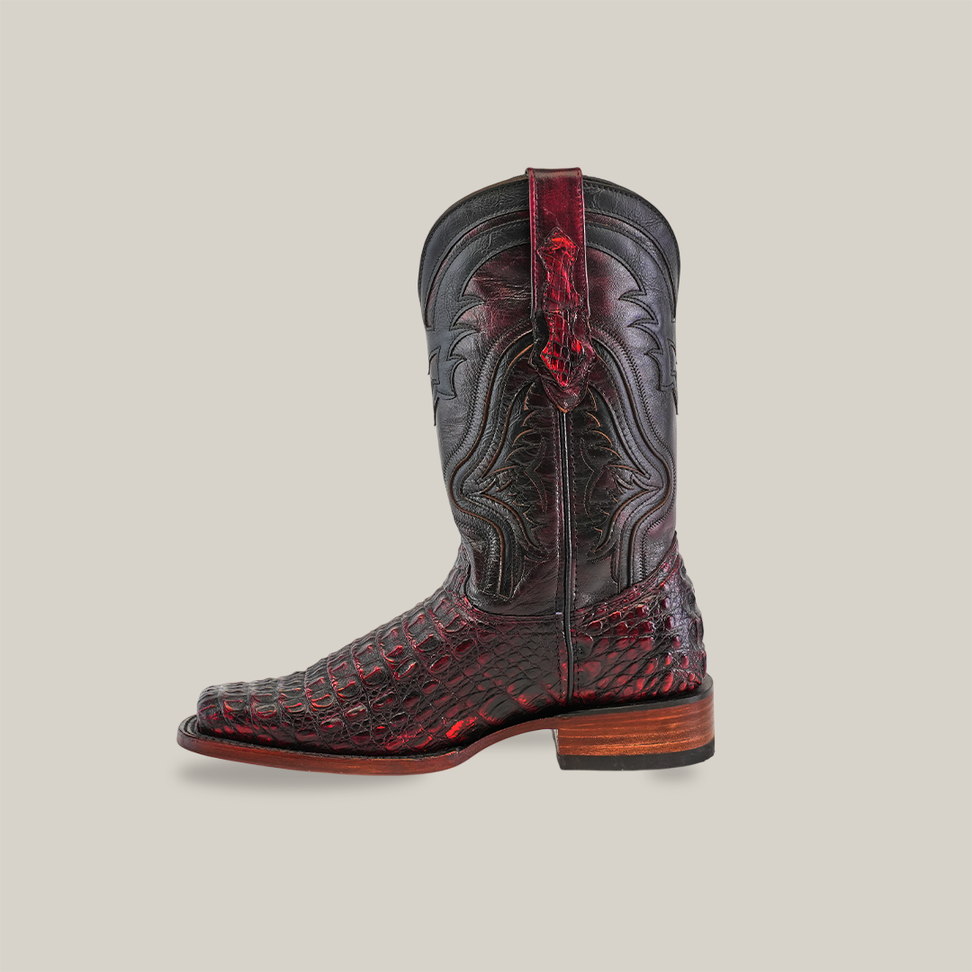 The Exotic Caiman Hornback - Black Cherry - Rodeo Toe is a single leather cowboy boot with intricate black and dark red detailing, ornate stitching, and a wooden heel that embodies the meeting of craftsmanship and tradition, set against a plain white background.