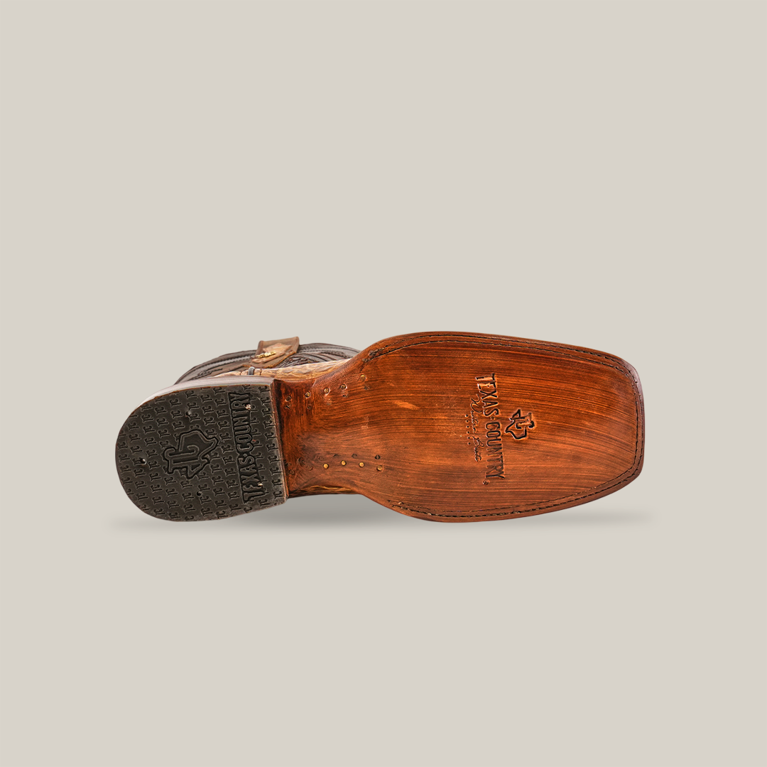 The bottom view of an Exotic Caiman Hornback boot displays a wooden sole and black rubber heel with TEK SQUARED logo. This antique rodeo toe cowboy boot, made from genuine tan caiman leather, is shown on its side against a plain white background.