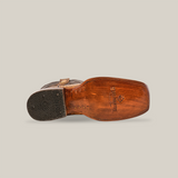 The bottom view of an Exotic Caiman Hornback boot displays a wooden sole and black rubber heel with TEK SQUARED logo. This antique rodeo toe cowboy boot, made from genuine tan caiman leather, is shown on its side against a plain white background.