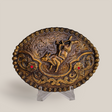 The Ruby Bull Rider Buckle is an ornate, oval bronze piece with an embossed horse amid intricate floral and swirl designs, echoing Texas-style buckles. Displayed on a clear stand against a simple backdrop, it exudes rustic charm and craftsmanship.